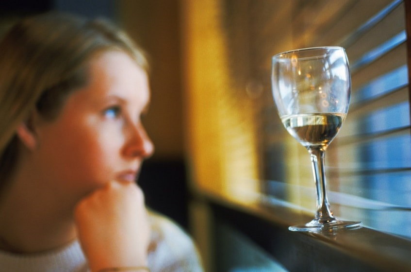 грустная женщина сидит возле окна с бокалом алкоголя